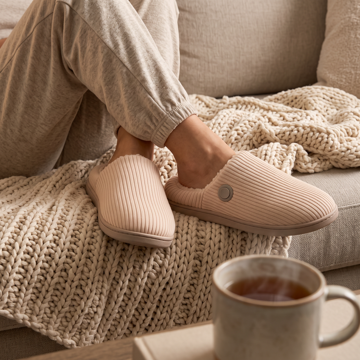 Woman relaxing on a sofa wearing blush Cozy Glow heated slippers